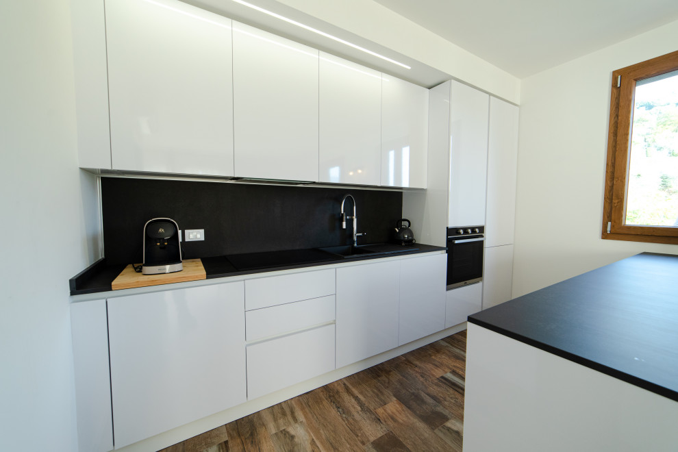 Photo of a mid-sized scandinavian galley eat-in kitchen in Milan with a drop-in sink, flat-panel cabinets, white cabinets, quartz benchtops, black splashback, engineered quartz splashback, black appliances, porcelain floors, with island, brown floor and black benchtop.