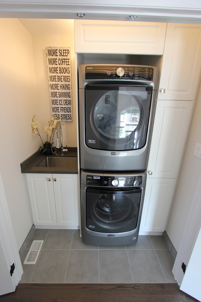 This is an example of a small contemporary single-wall laundry cupboard in Toronto with an undermount sink, raised-panel cabinets, white cabinets, white walls, ceramic floors, a stacked washer and dryer and grey floor.