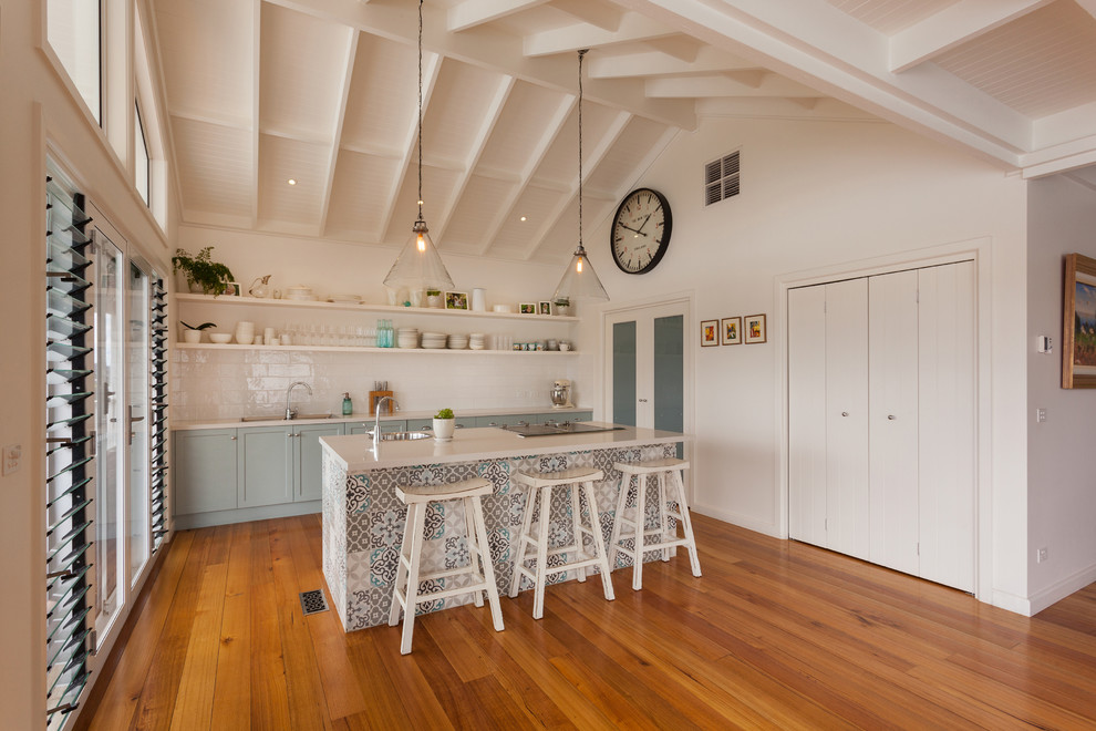 Beach style galley open plan kitchen with a drop-in sink, shaker cabinets, blue cabinets, white splashback, subway tile splashback, panelled appliances, medium hardwood floors, with island, brown floor and white benchtop.