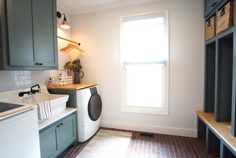 Mid-sized country galley dedicated laundry room in Philadelphia with a farmhouse sink, shaker cabinets, green cabinets, quartz benchtops, grey walls, brick floors, a side-by-side washer and dryer, multi-coloured floor and white benchtop.