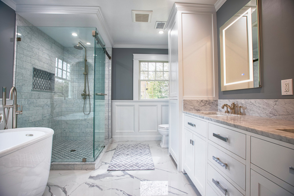 Inspiration for a large transitional master bathroom in New York with shaker cabinets, white cabinets, a freestanding tub, an alcove shower, a one-piece toilet, white tile, marble, grey walls, porcelain floors, an undermount sink, marble benchtops, white floor, a sliding shower screen, white benchtops, a double vanity, a freestanding vanity and panelled walls.