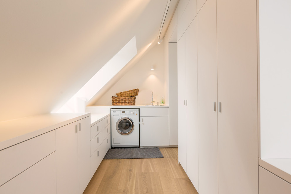 This is an example of a mid-sized modern u-shaped dedicated laundry room in Munich with a drop-in sink, flat-panel cabinets, white cabinets, white walls, light hardwood floors and a side-by-side washer and dryer.