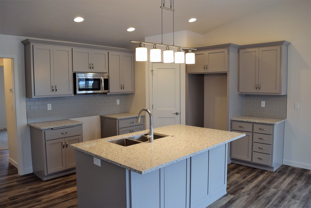 Large l-shaped medium tone wood floor open concept kitchen photo with a double-bowl sink, raised-panel cabinets, gray cabinets, granite countertops, gray backsplash, ceramic backsplash and an island