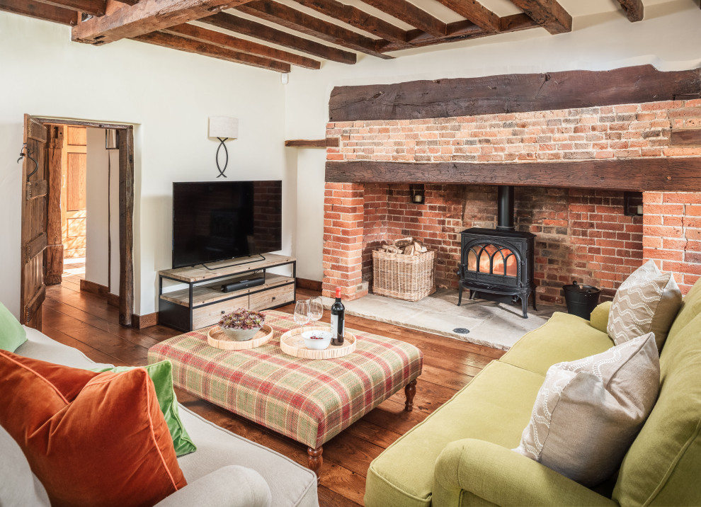 Photo of a large farmhouse enclosed living room in Surrey with white walls, medium hardwood flooring, a standard fireplace, a brick fireplace surround, a freestanding tv and exposed beams.