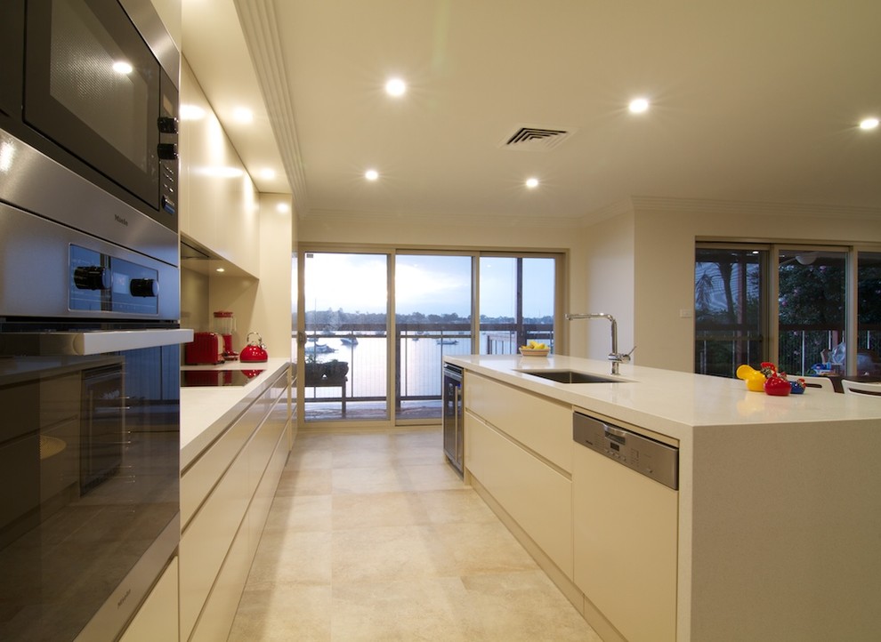 This is an example of a mid-sized contemporary galley open plan kitchen in Sydney with with island, an undermount sink, flat-panel cabinets, beige cabinets, quartz benchtops, brown splashback, glass sheet splashback, stainless steel appliances, travertine floors, beige floor and beige benchtop.