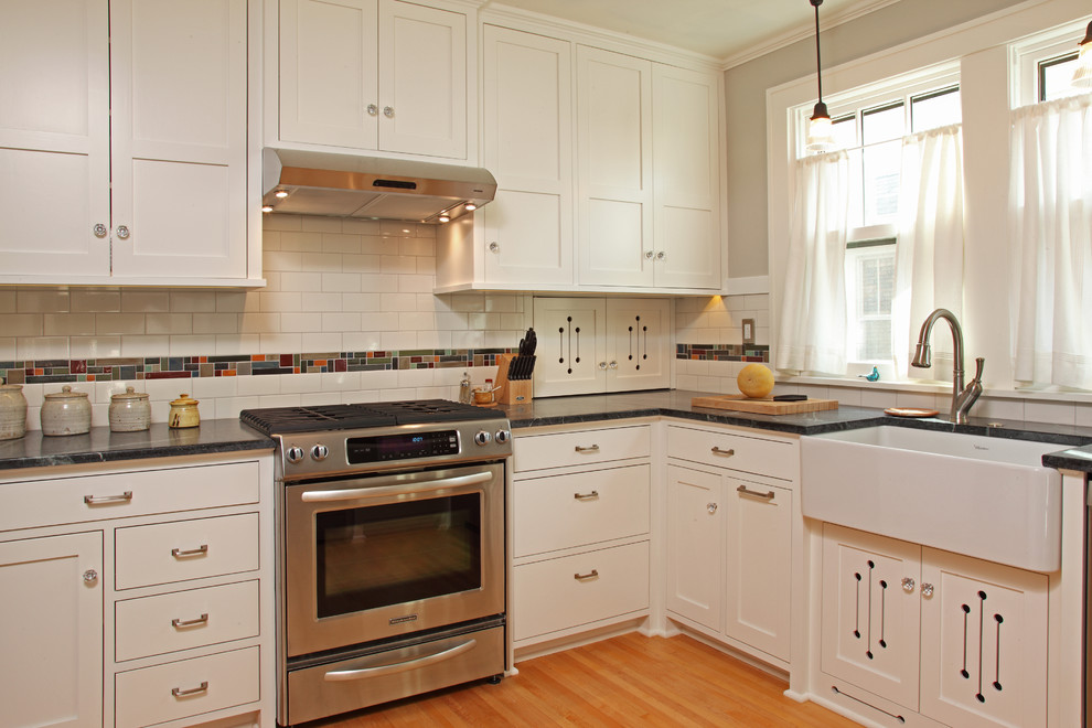 Small arts and crafts l-shaped eat-in kitchen in Minneapolis with a farmhouse sink, recessed-panel cabinets, white cabinets, multi-coloured splashback, mosaic tile splashback, stainless steel appliances, light hardwood floors, no island, soapstone benchtops and beige floor.