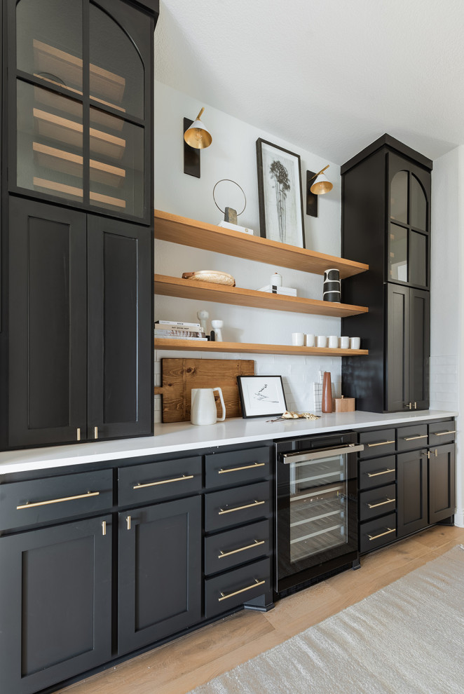 Inspiration for a mid-sized modern galley home bar in Dallas with no sink, shaker cabinets, black cabinets, white splashback, light hardwood floors and white benchtop.