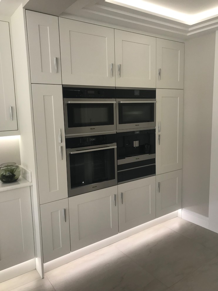 This is an example of a large contemporary open plan kitchen in Hertfordshire with a built-in sink, shaker cabinets, white cabinets, quartz worktops, white splashback, stone slab splashback, integrated appliances, porcelain flooring, an island, grey floors and white worktops.