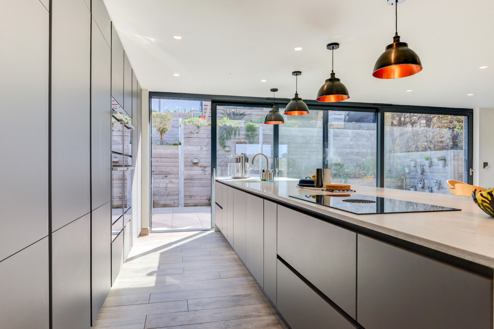 This is an example of an expansive modern single-wall open plan kitchen in Sussex with an integrated sink, flat-panel cabinets, grey cabinets, solid surface benchtops, black appliances, light hardwood floors, with island, brown floor and grey benchtop.
