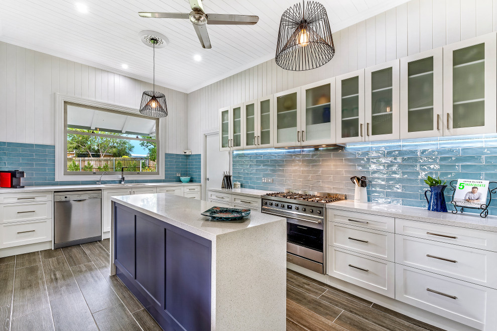 This is an example of a beach style l-shaped kitchen in Brisbane with an undermount sink, glass-front cabinets, white cabinets, blue splashback, subway tile splashback, stainless steel appliances, with island, brown floor, grey benchtop and timber.