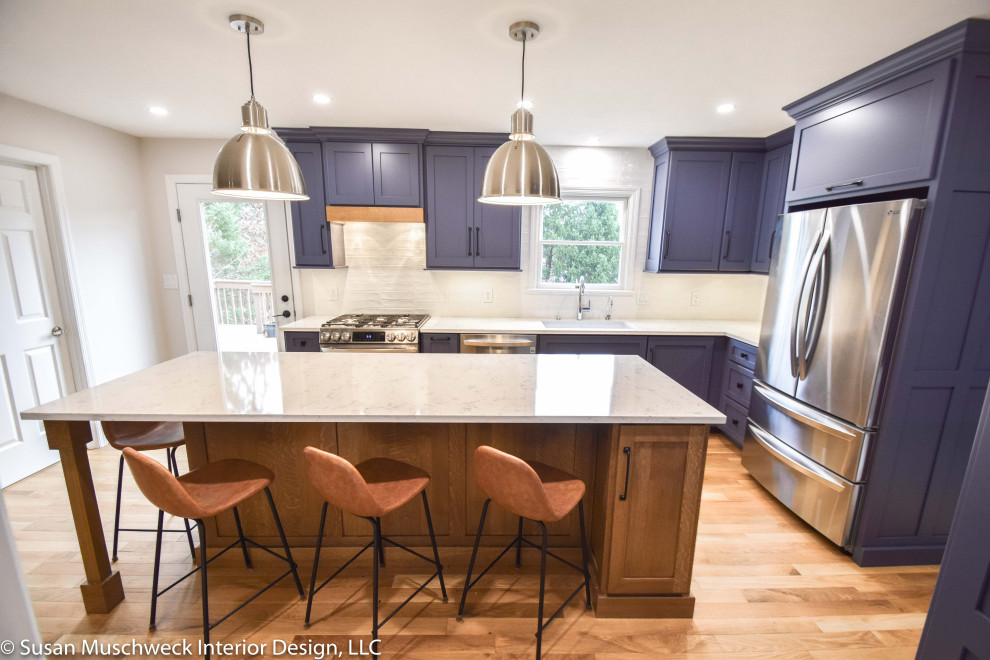 Example of a mid-sized transitional l-shaped light wood floor enclosed kitchen design with an undermount sink, shaker cabinets, blue cabinets, quartz countertops, white backsplash, ceramic backsplash, stainless steel appliances, an island and white countertops