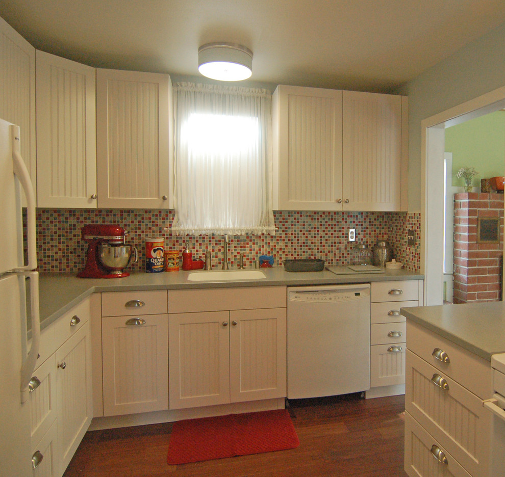 This is an example of a small beach style l-shaped separate kitchen in San Luis Obispo with an integrated sink, beaded inset cabinets, white cabinets, solid surface benchtops, multi-coloured splashback, glass tile splashback, white appliances and dark hardwood floors.