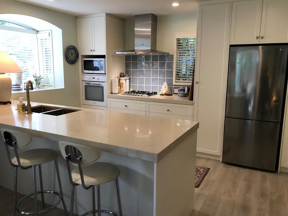 This is an example of a mid-sized traditional galley open plan kitchen in Christchurch with a double-bowl sink, shaker cabinets, white cabinets, quartz benchtops, blue splashback, glass tile splashback, white appliances, light hardwood floors, a peninsula and multi-coloured floor.