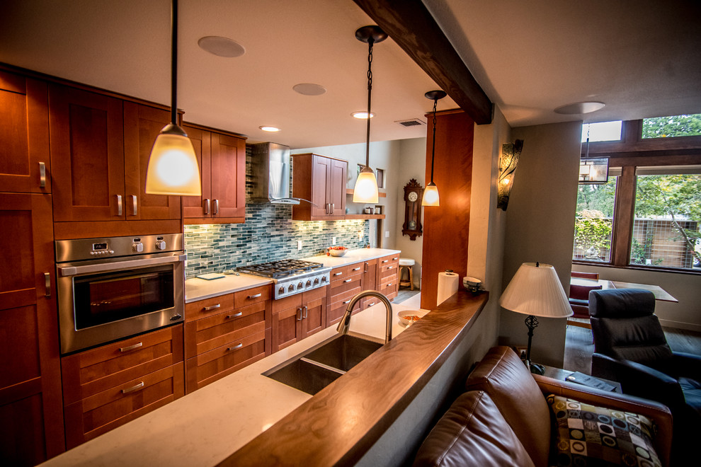 This is an example of a large contemporary galley eat-in kitchen in Portland with a double-bowl sink, shaker cabinets, quartz benchtops, stainless steel appliances, brown cabinets, green splashback, glass tile splashback, laminate floors, brown floor, white benchtop and a peninsula.