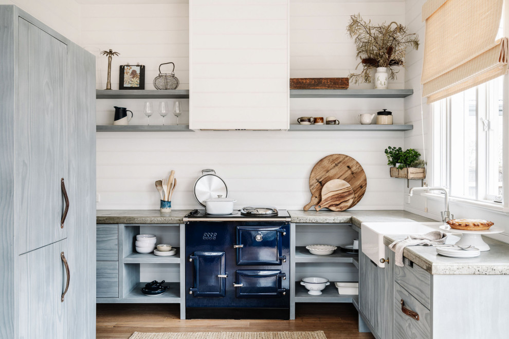 Photo of a mid-sized beach style u-shaped eat-in kitchen in Melbourne with a farmhouse sink, open cabinets, distressed cabinets, concrete benchtops, white splashback, shiplap splashback, coloured appliances, light hardwood floors, brown floor and grey benchtop.
