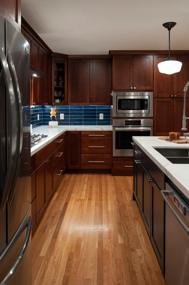 This is an example of a large transitional l-shaped eat-in kitchen in San Francisco with a double-bowl sink, shaker cabinets, brown cabinets, blue splashback, glass tile splashback, stainless steel appliances, light hardwood floors, with island, yellow floor and white benchtop.