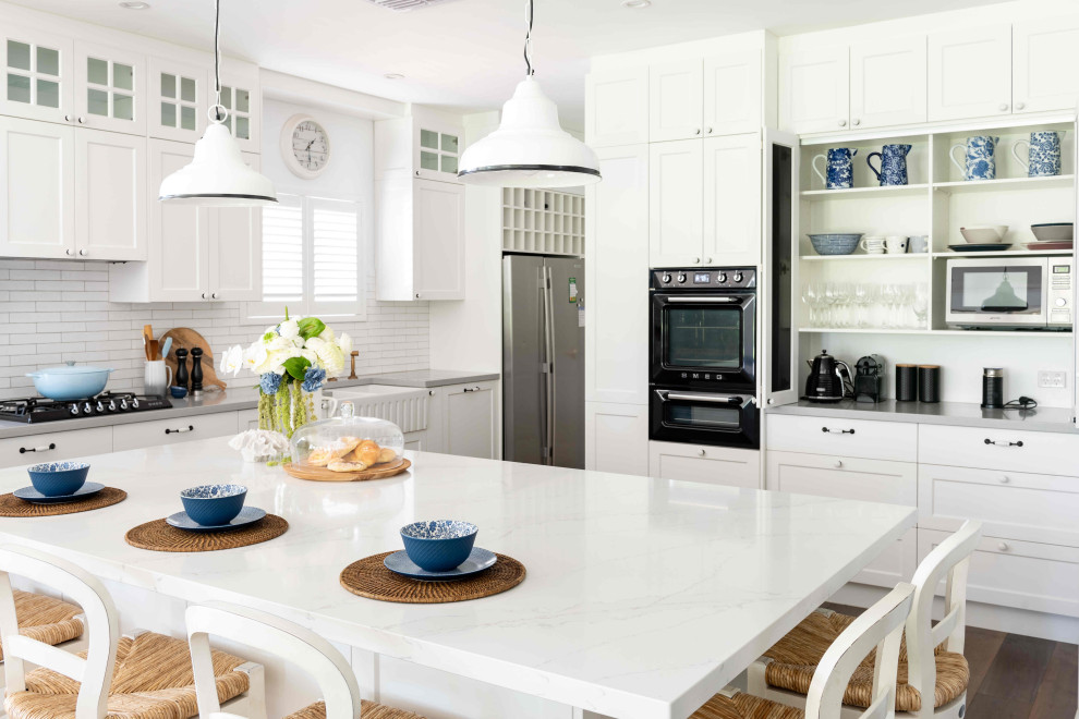 Photo of a large beach style u-shaped open plan kitchen in Perth with a farmhouse sink, raised-panel cabinets, white cabinets, marble benchtops, white splashback, subway tile splashback, black appliances, dark hardwood floors, with island and grey benchtop.