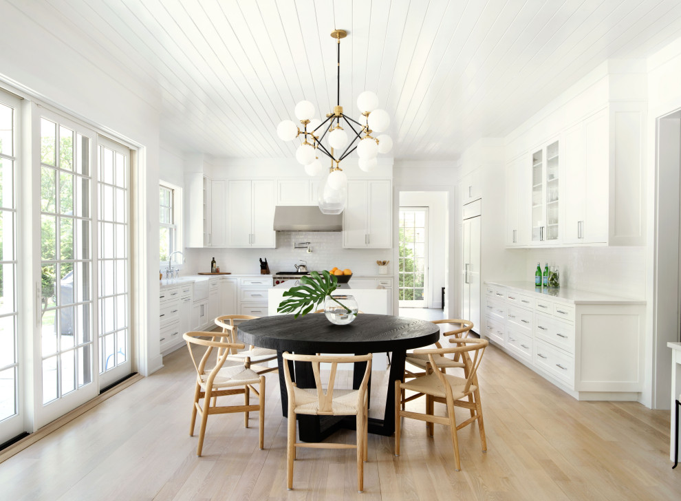 Beach style u-shaped eat-in kitchen in New York with a farmhouse sink, shaker cabinets, white cabinets, white splashback, subway tile splashback, light hardwood floors, with island, beige floor, white benchtop and panelled appliances.