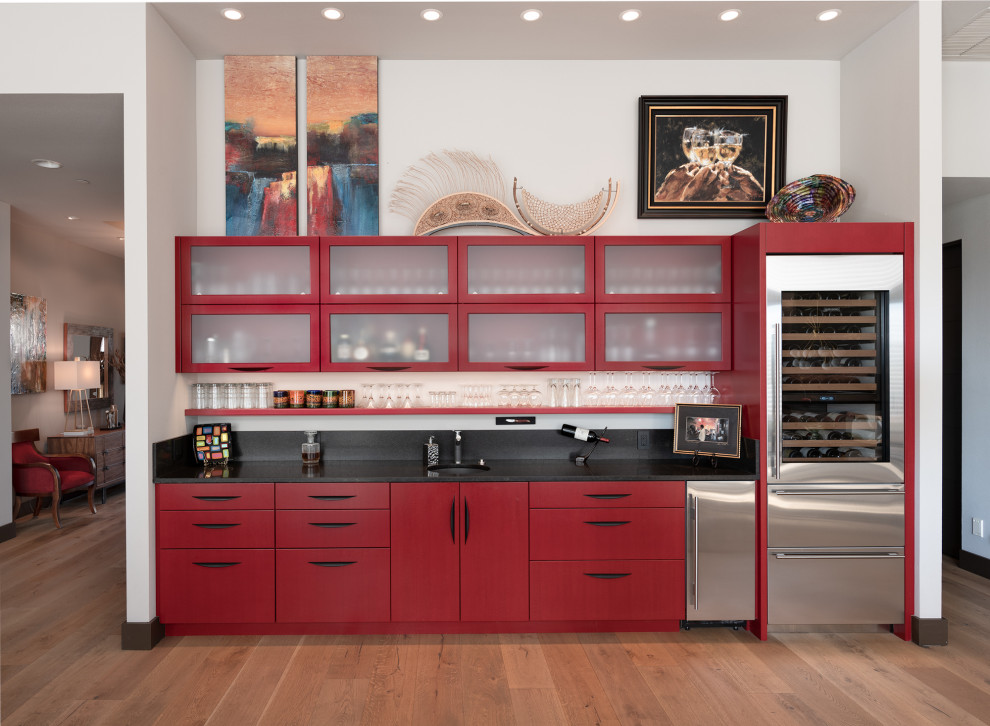 Contemporary single-wall wet bar in Portland with an undermount sink, flat-panel cabinets, red cabinets, medium hardwood floors, brown floor and black benchtop.