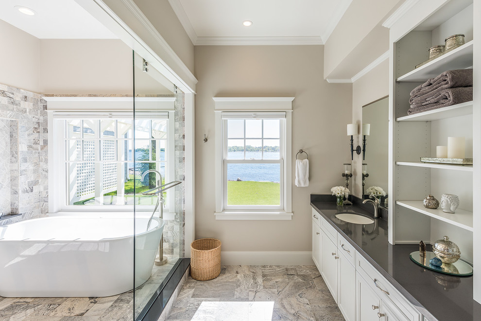 This is an example of a large traditional master bathroom in Minneapolis with flat-panel cabinets, white cabinets, a freestanding tub, an open shower, multi-coloured tile, stone tile, an undermount sink, beige walls, porcelain floors, solid surface benchtops and grey benchtops.