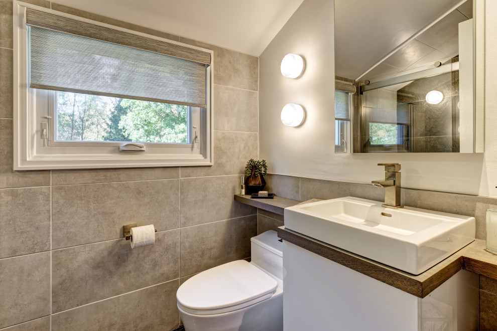 Bild på ett litet funkis brun brunt badrum med dusch, med släta luckor, vita skåp, en dusch i en alkov, en toalettstol med hel cisternkåpa, grå kakel, porslinskakel, grå väggar, mosaikgolv, ett avlångt handfat, träbänkskiva, brunt golv och dusch med skjutdörr