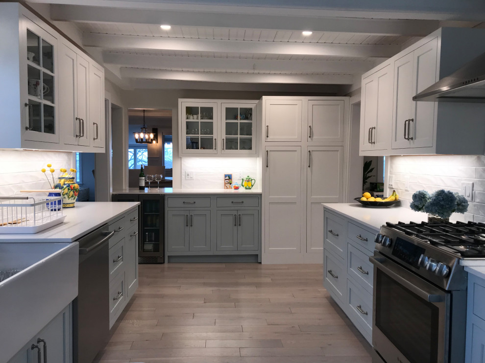 Photo of a mid-sized beach style u-shaped separate kitchen in Boston with a farmhouse sink, shaker cabinets, blue cabinets, quartz benchtops, white splashback, subway tile splashback, stainless steel appliances, light hardwood floors, a peninsula, beige floor, white benchtop and vaulted.