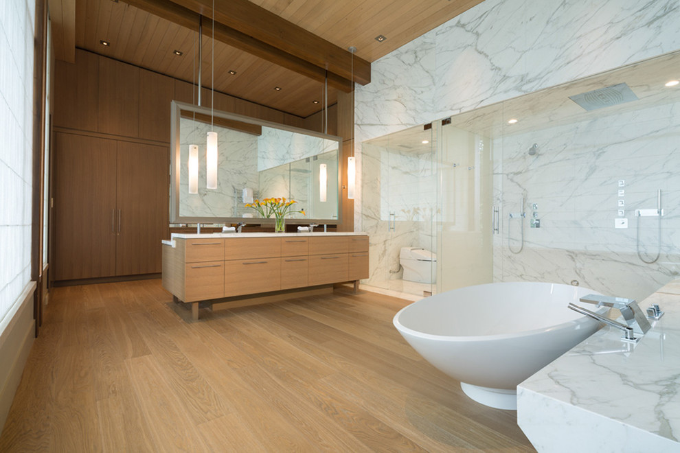 Expansive contemporary master bathroom in Vancouver with flat-panel cabinets, light wood cabinets, a freestanding tub, a double shower, a one-piece toilet, stone slab, white walls, light hardwood floors, a wall-mount sink, marble benchtops, white tile and a hinged shower door.