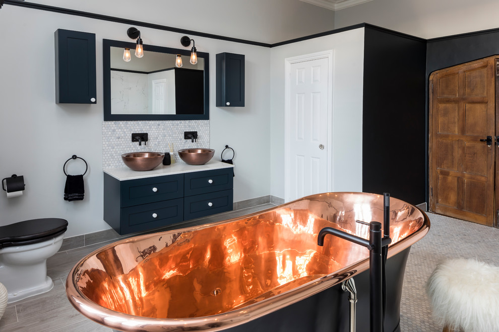 Inspiration for a large transitional master bathroom in Surrey with shaker cabinets, blue cabinets, a freestanding tub, a two-piece toilet, gray tile, ceramic tile, white walls, ceramic floors, a wall-mount sink, solid surface benchtops and white benchtops.