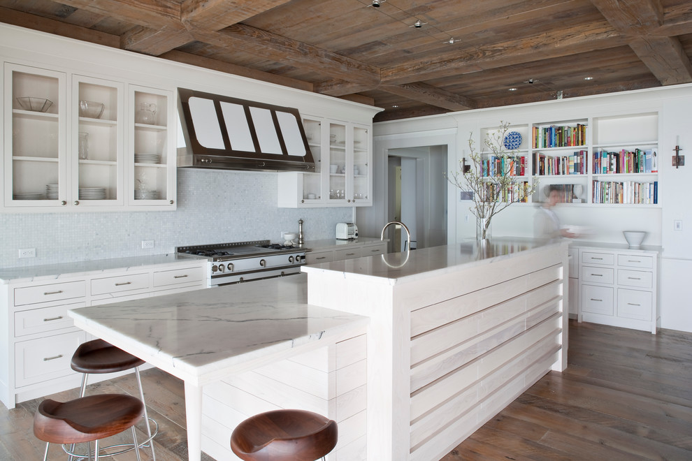 Photo of a beach style galley kitchen in New York with glass-front cabinets, white cabinets, white splashback, mosaic tile splashback, white appliances, dark hardwood floors and with island.