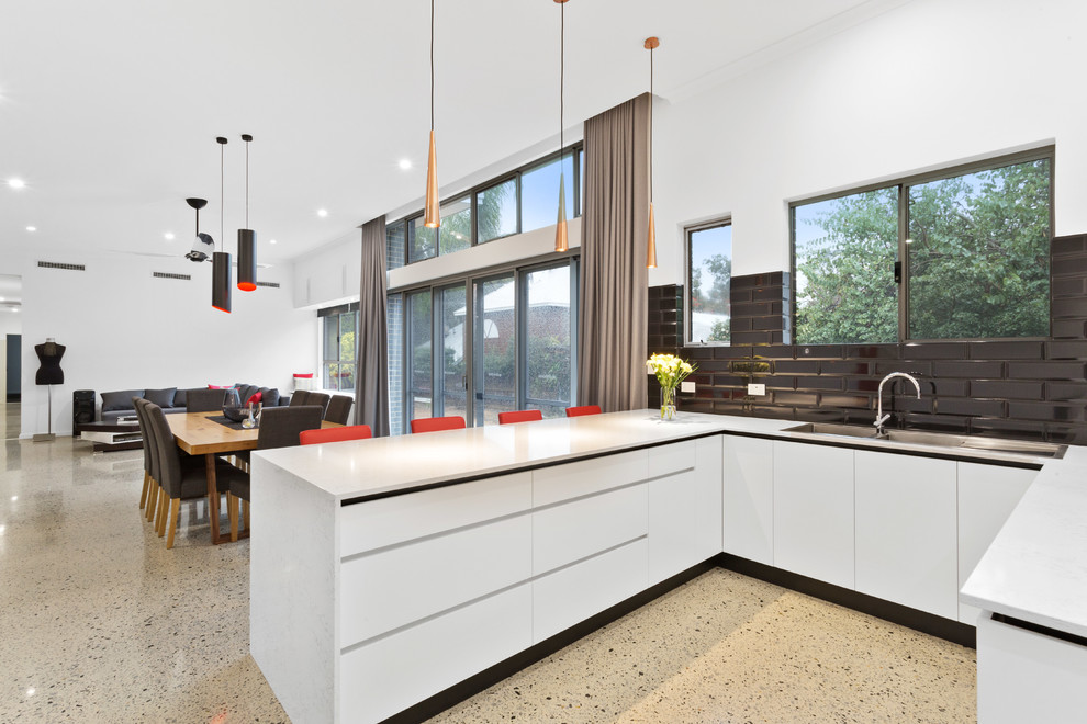This is an example of an expansive modern l-shaped eat-in kitchen in Perth with a drop-in sink, flat-panel cabinets, black appliances, concrete floors, no island, grey floor, white cabinets, solid surface benchtops, black splashback, subway tile splashback and white benchtop.