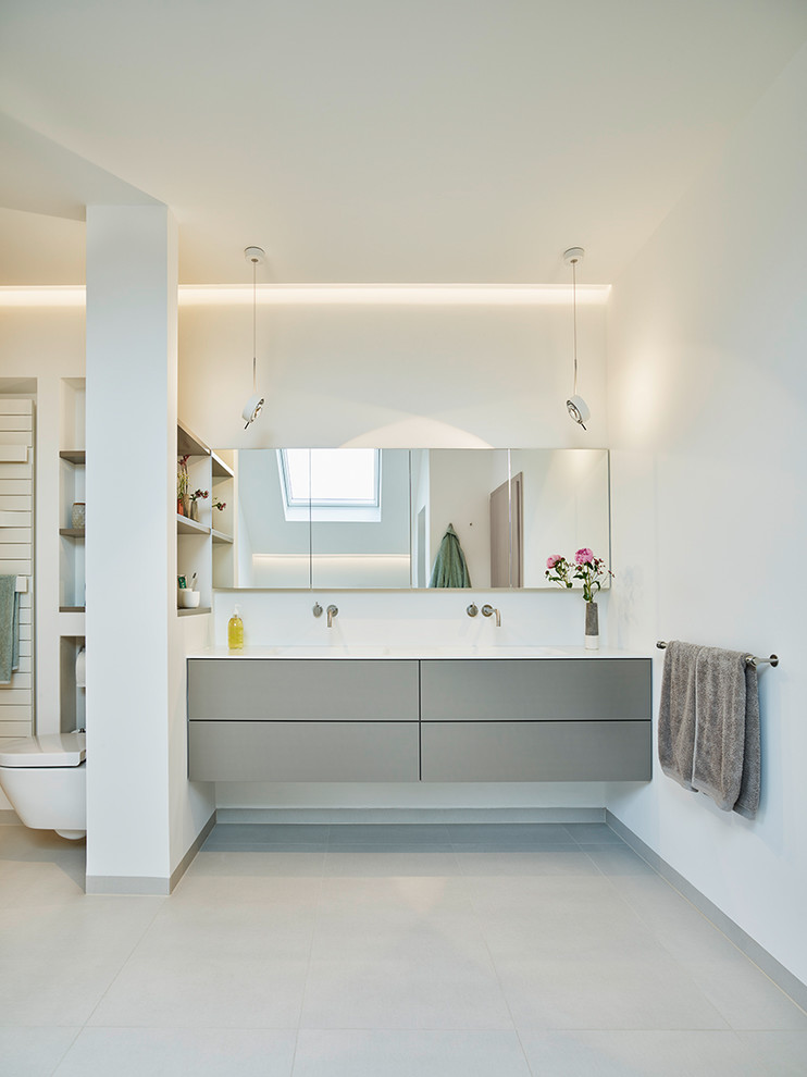 This is an example of a mid-sized contemporary bathroom in Frankfurt with flat-panel cabinets, grey cabinets, a wall-mount toilet, white walls, an integrated sink, grey floor and white benchtops.