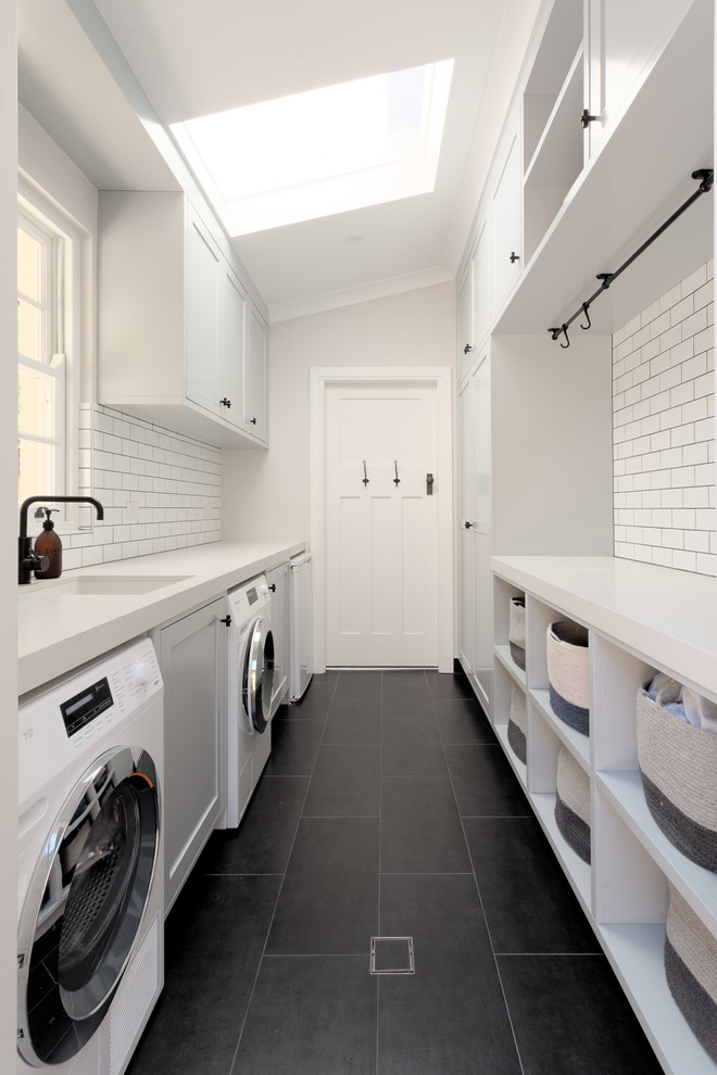 Photo of a small beach style galley dedicated laundry room in Sydney with flat-panel cabinets, white cabinets, white walls, a side-by-side washer and dryer, black floor and an undermount sink.