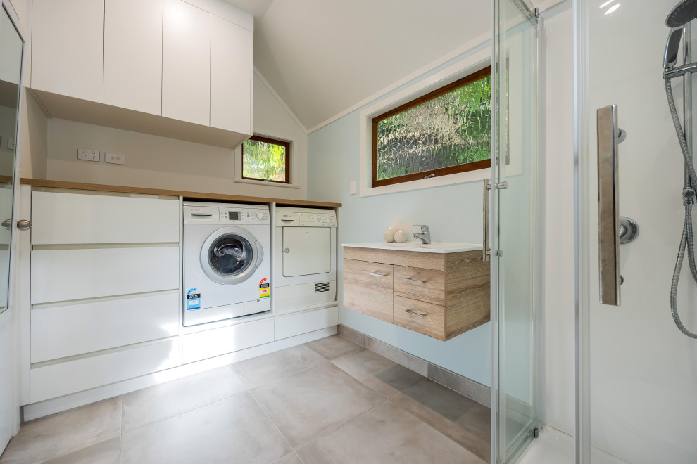 This is an example of a small modern single-wall utility room in Auckland with flat-panel cabinets, white cabinets, wood benchtops, blue splashback, white walls, porcelain floors, a side-by-side washer and dryer, grey floor and brown benchtop.