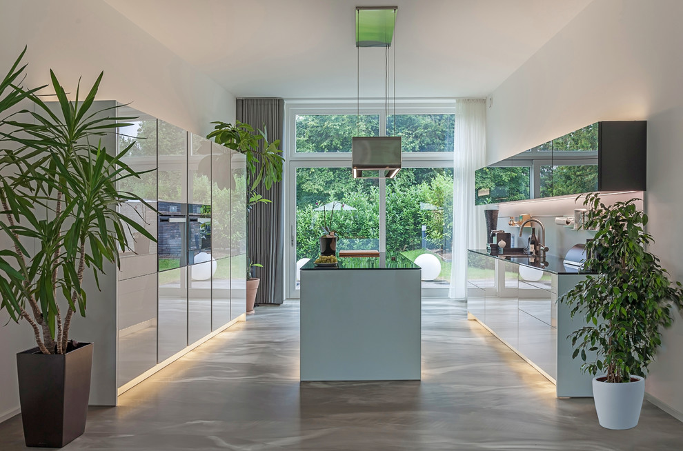 Expansive contemporary galley open plan kitchen in Dusseldorf with a drop-in sink, flat-panel cabinets, white cabinets, grey splashback, black appliances, with island, grey floor, solid surface benchtops, glass sheet splashback, concrete floors and black benchtop.