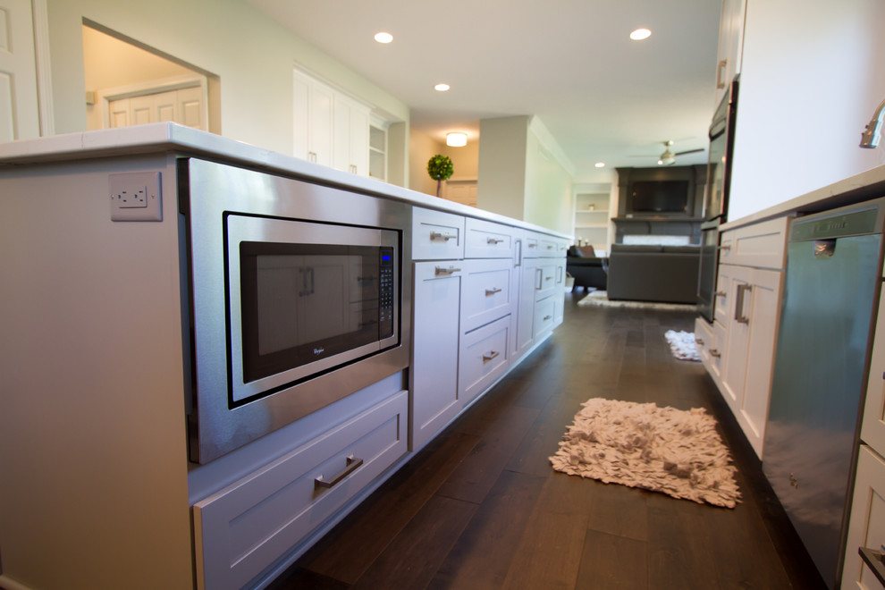 Example of a transitional single-wall dark wood floor and brown floor open concept kitchen design with an undermount sink, recessed-panel cabinets, white cabinets, quartzite countertops, gray backsplash, marble backsplash, stainless steel appliances and an island