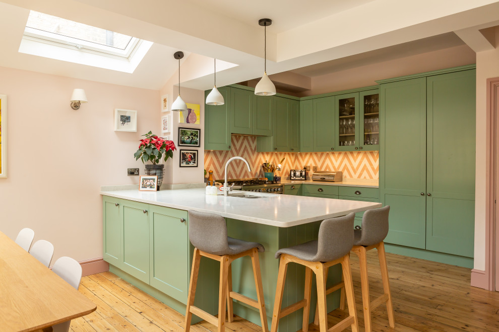 This is an example of a transitional u-shaped kitchen in Essex with an undermount sink, shaker cabinets, green cabinets, orange splashback, panelled appliances, medium hardwood floors, a peninsula, brown floor and white benchtop.