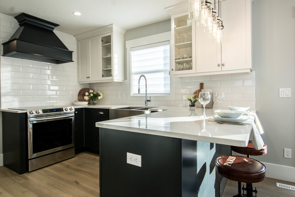 Open concept kitchen - mid-sized transitional u-shaped medium tone wood floor and brown floor open concept kitchen idea in Calgary with a farmhouse sink, recessed-panel cabinets, black cabinets, quartz countertops, white backsplash, subway tile backsplash, stainless steel appliances and a peninsula