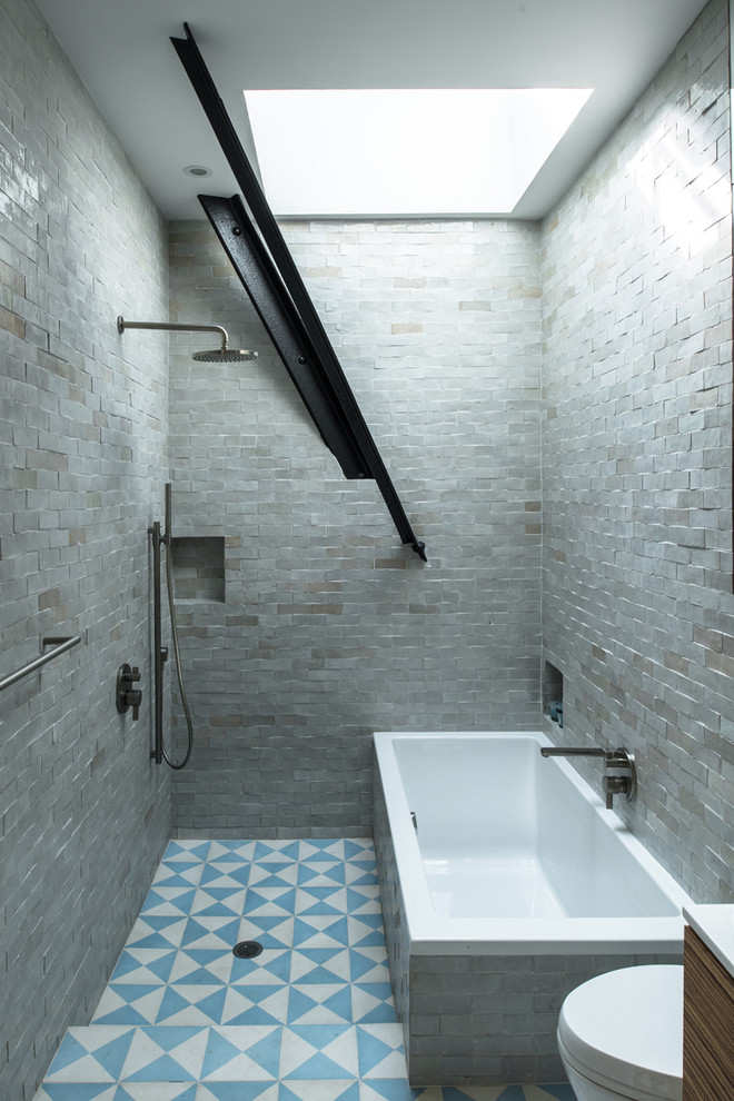 Mid-sized industrial master bathroom in New York with a drop-in tub, an open shower, an open shower, gray tile, stone tile, flat-panel cabinets, medium wood cabinets, a drop-in sink, solid surface benchtops, a wall-mount toilet, grey walls and ceramic floors.