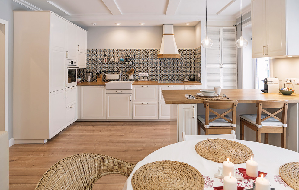 Inspiration for a scandinavian l-shaped open plan kitchen in Yekaterinburg with a farmhouse sink, white cabinets, multi-coloured splashback, white appliances, light hardwood floors and a peninsula.