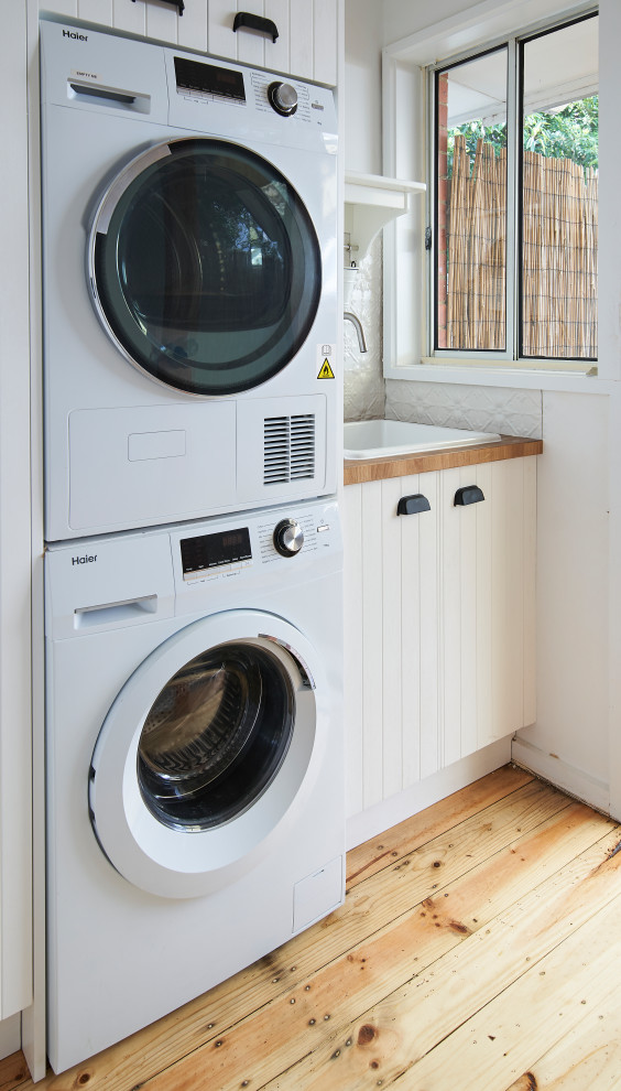 Photo of a small modern single-wall dedicated laundry room in Melbourne with a farmhouse sink, shaker cabinets, white cabinets, wood benchtops, white splashback, white walls, medium hardwood floors, a stacked washer and dryer, beige floor and brown benchtop.