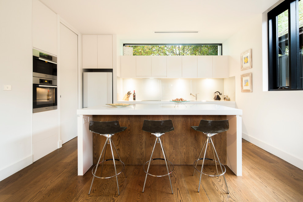 Design ideas for a mid-sized modern galley kitchen in Melbourne with an undermount sink, flat-panel cabinets, white cabinets, quartz benchtops, white splashback, glass sheet splashback, stainless steel appliances, medium hardwood floors and with island.