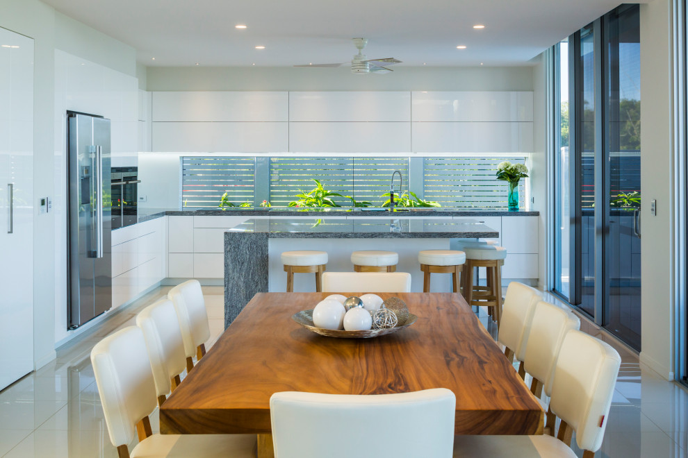 This is an example of a contemporary l-shaped eat-in kitchen in Cairns with an undermount sink, granite benchtops, white splashback, glass sheet splashback, stainless steel appliances, porcelain floors, with island, white floor, grey benchtop, flat-panel cabinets and white cabinets.