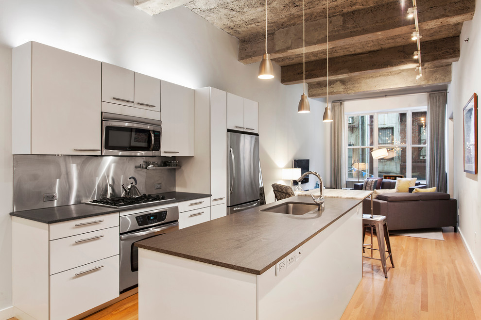 Photo of an industrial open plan kitchen in New York with flat-panel cabinets, white cabinets, metallic splashback, stainless steel appliances, light hardwood floors and with island.