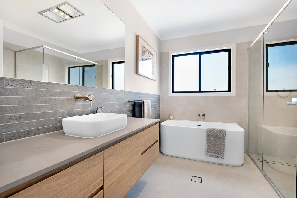 Modern inredning av ett grå grått badrum, med släta luckor, skåp i ljust trä, ett fristående badkar, en kantlös dusch, grå kakel, grå väggar, ett fristående handfat, grått golv och dusch med gångjärnsdörr