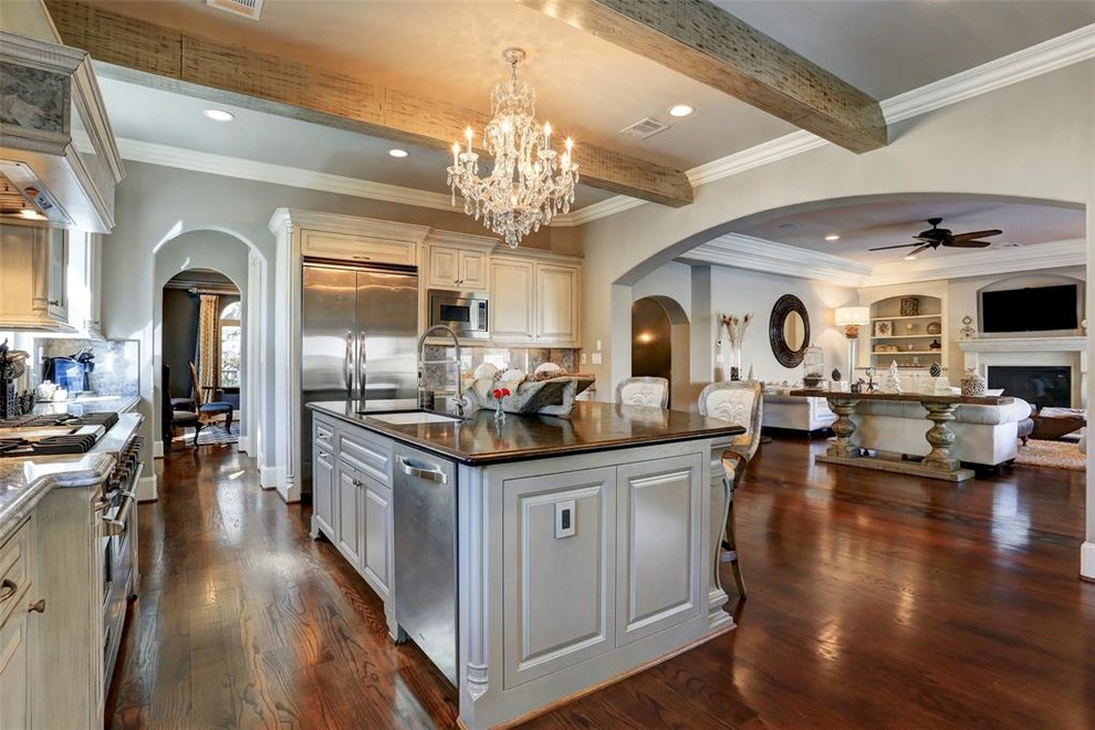 Photo of a large mediterranean l-shaped open plan kitchen in Houston with raised-panel cabinets, granite benchtops, with island, black benchtop, an undermount sink, distressed cabinets, grey splashback, marble splashback, stainless steel appliances, dark hardwood floors and brown floor.