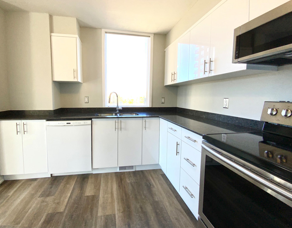 Small contemporary l-shaped kitchen/diner in Calgary with a double-bowl sink, flat-panel cabinets, white cabinets, granite worktops, black splashback, granite splashback, stainless steel appliances, laminate floors, brown floors and black worktops.