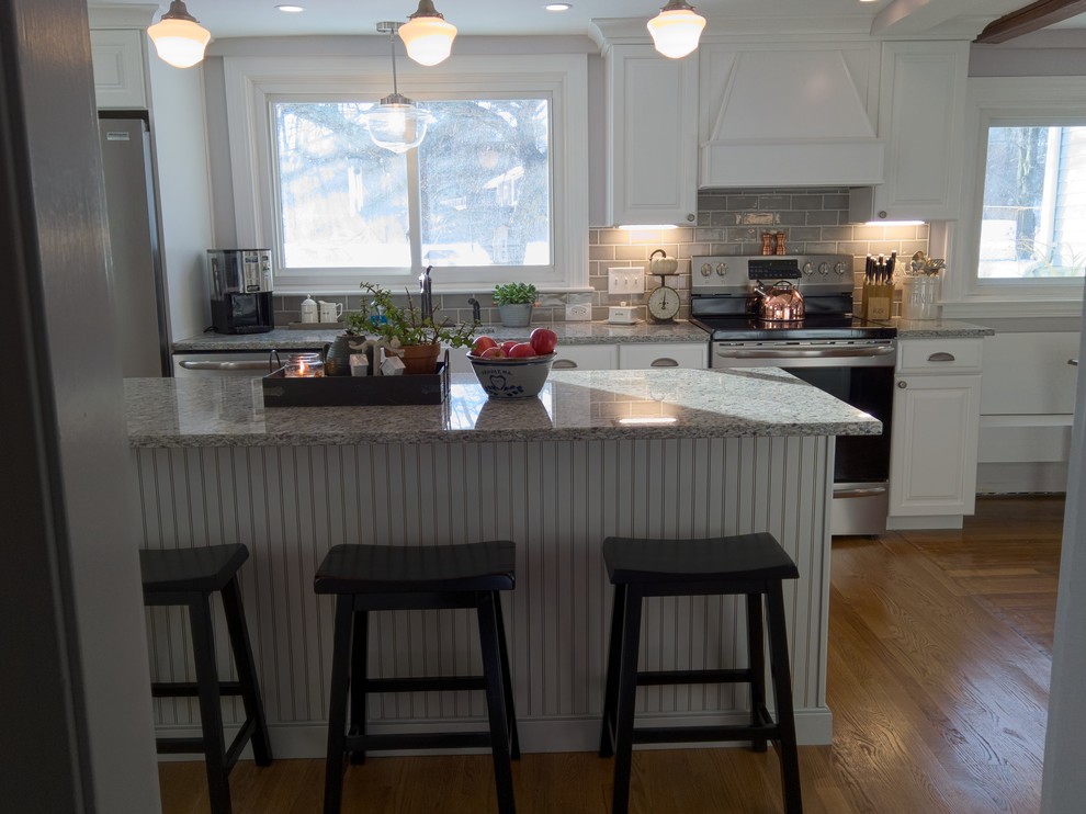 Design ideas for a mid-sized country kitchen in Boston with a farmhouse sink, raised-panel cabinets, white cabinets, beige splashback, subway tile splashback, stainless steel appliances, light hardwood floors, with island and grey benchtop.