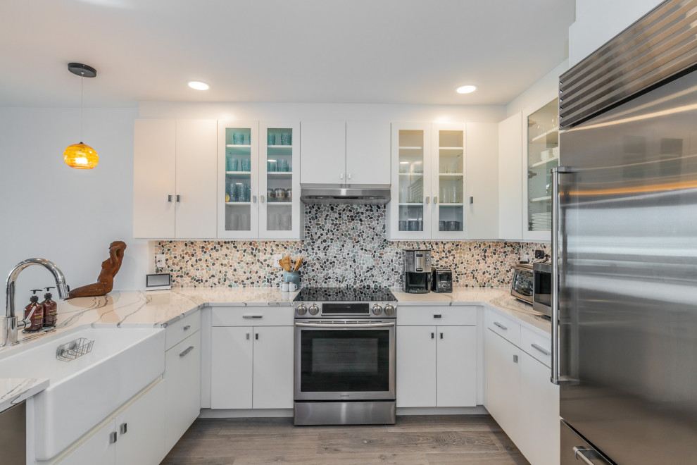 Photo of a large beach style u-shaped eat-in kitchen in Tampa with a farmhouse sink, flat-panel cabinets, white cabinets, quartzite benchtops, multi-coloured splashback, mosaic tile splashback, stainless steel appliances, porcelain floors, a peninsula, grey floor and white benchtop.