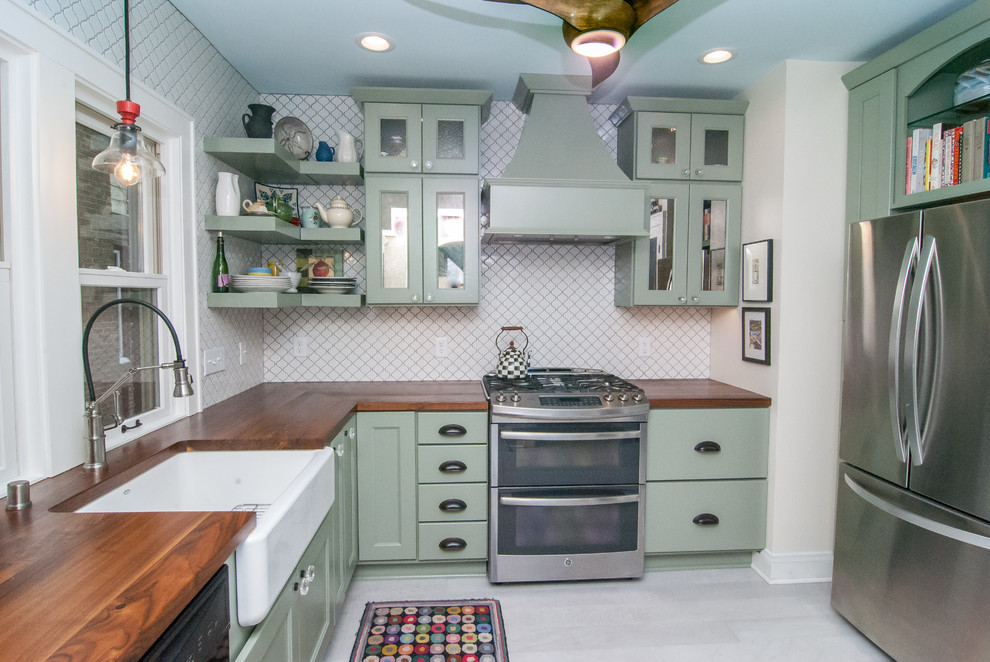This is an example of an eclectic l-shaped kitchen in Milwaukee with a farmhouse sink, shaker cabinets, green cabinets, wood benchtops, white splashback, porcelain splashback, stainless steel appliances, porcelain floors, grey floor and brown benchtop.