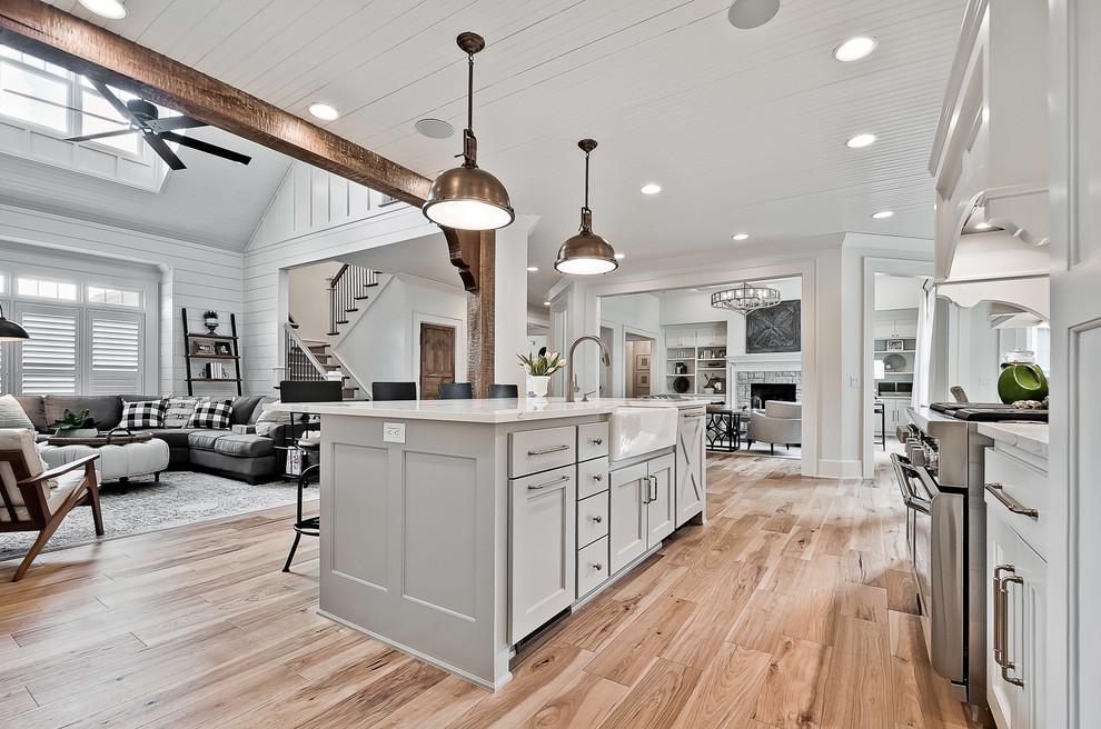 This is an example of a large arts and crafts open plan kitchen in Other with a farmhouse sink, raised-panel cabinets, white cabinets, quartz benchtops, white splashback, stone tile splashback, stainless steel appliances, light hardwood floors, with island, brown floor and white benchtop.